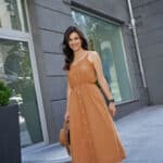 caucasian woman wearing long romantic dress holding hat, looking at camera and smiling while standing on the city street