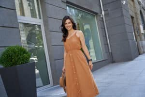 caucasian woman wearing long romantic dress holding hat, looking at camera and smiling while standing on the city street